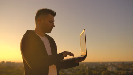 Hacker-using-laptop-on-rooftop-with-city-view-and-forex-chart.-Hacking-and-stats-concept.-A-man-at-sunset-in-slow-motion-writing-software-code-on-a-laptop